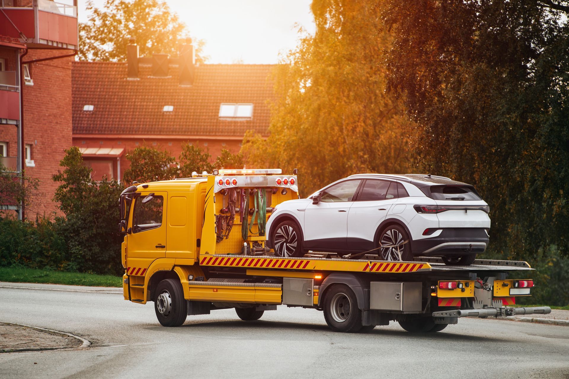 Yellow tow truck carrying a white SUV on a flatbed at sunset on a street
