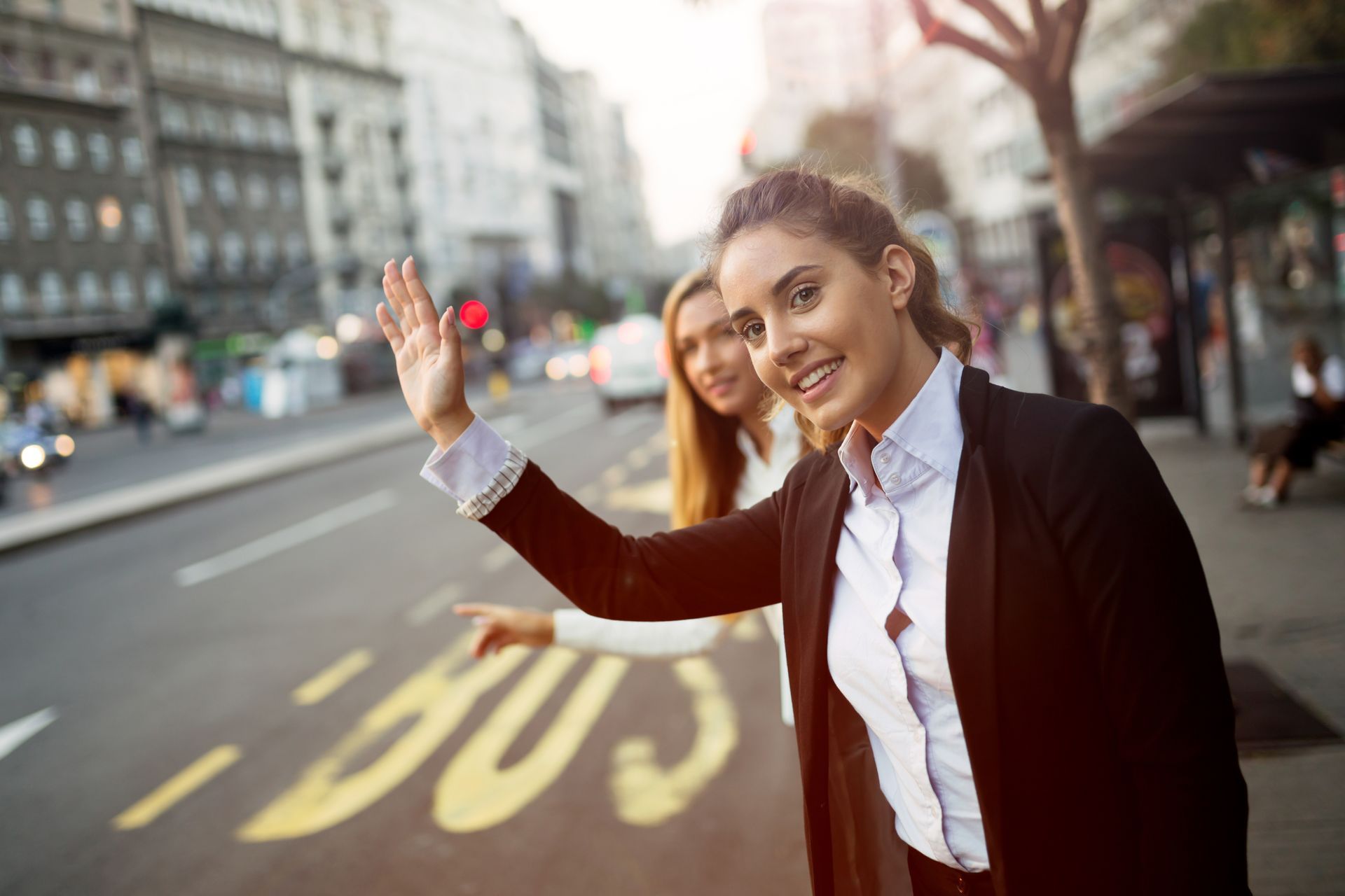 Two people waving on a city street, one in a black blazer and white shirt, with buildings in the background
