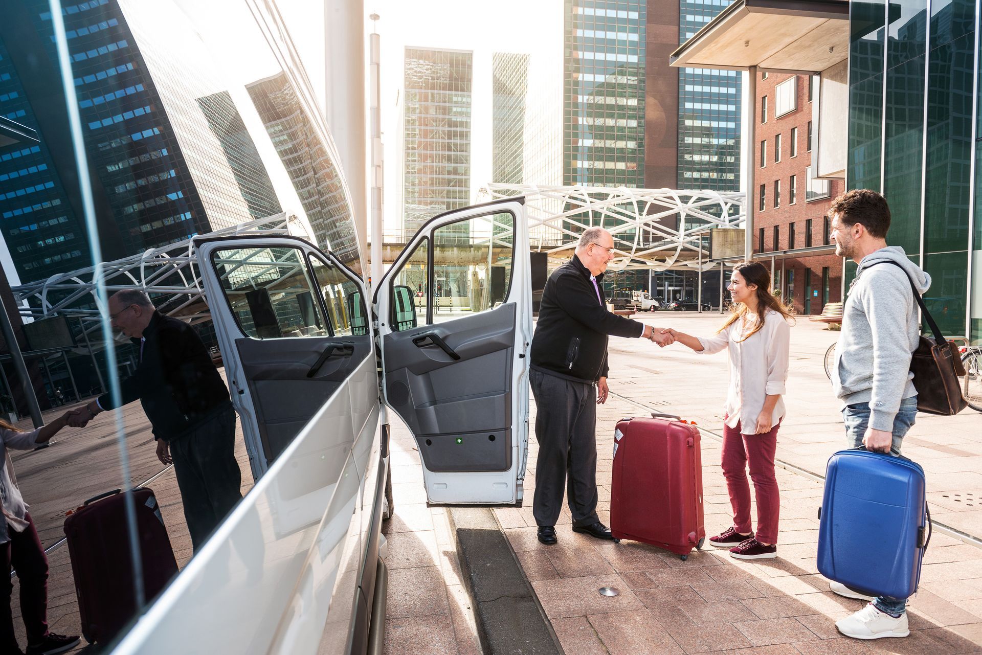Travelers with rolling suitcases meet a driver beside a car on a city sidewalk.