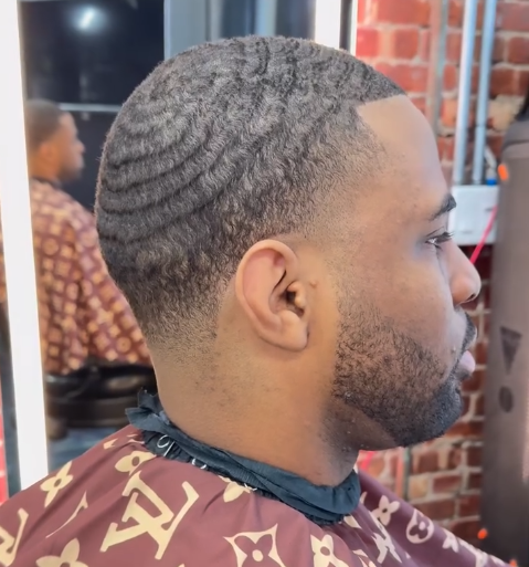 Side-profile of a man with a short wave haircut in a barber chair, wearing a salon cape.