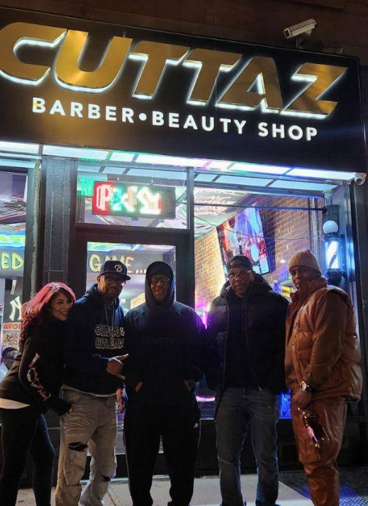 Five people posing outside CUTTAZ Barber & Beauty Shop at night, neon signs glowing.
