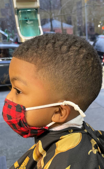 Child in profile wearing a red plaid face mask and black-and-gold cape indoors.