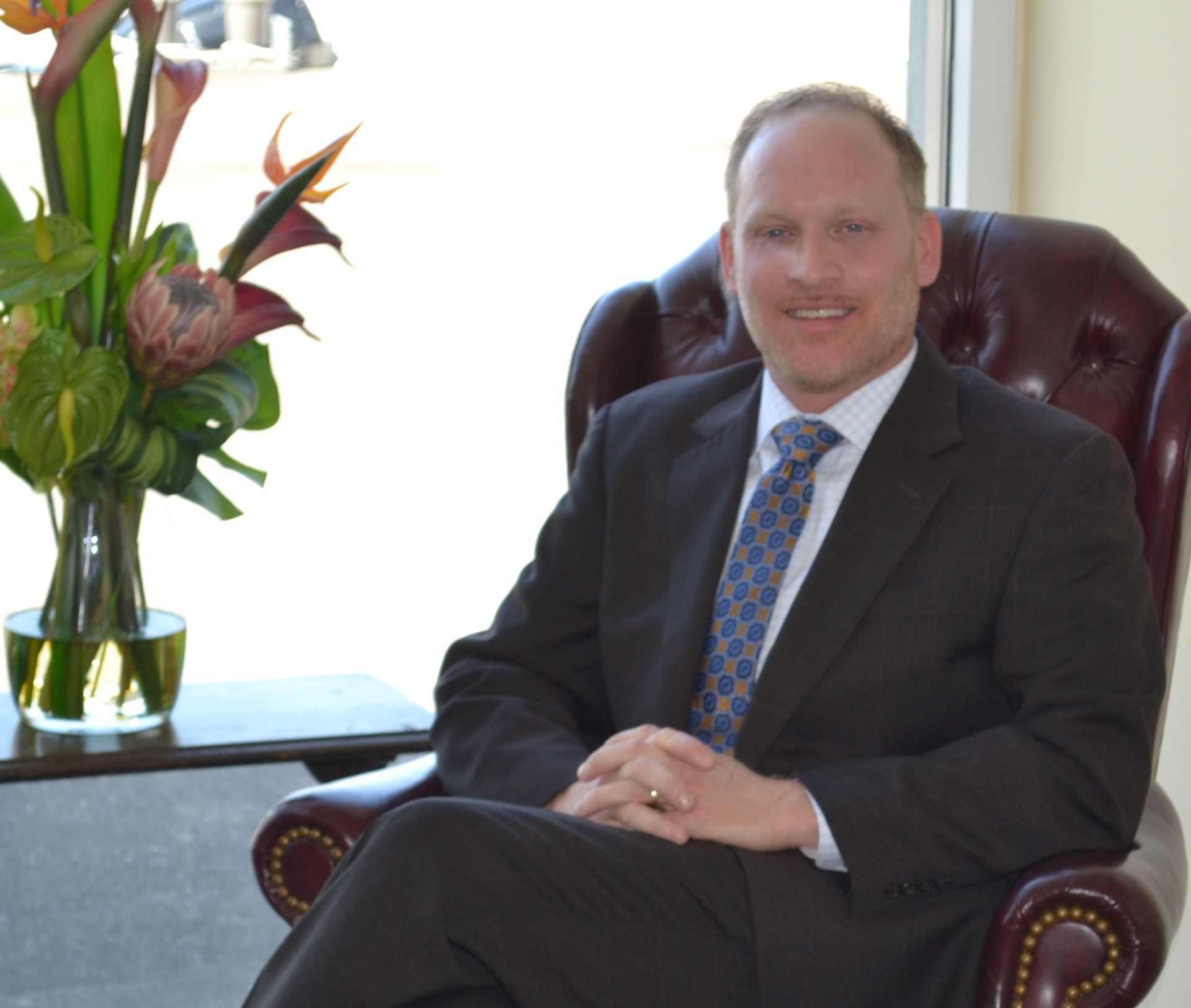 Man in suit, seated, smiling; next to flowers, window in background.