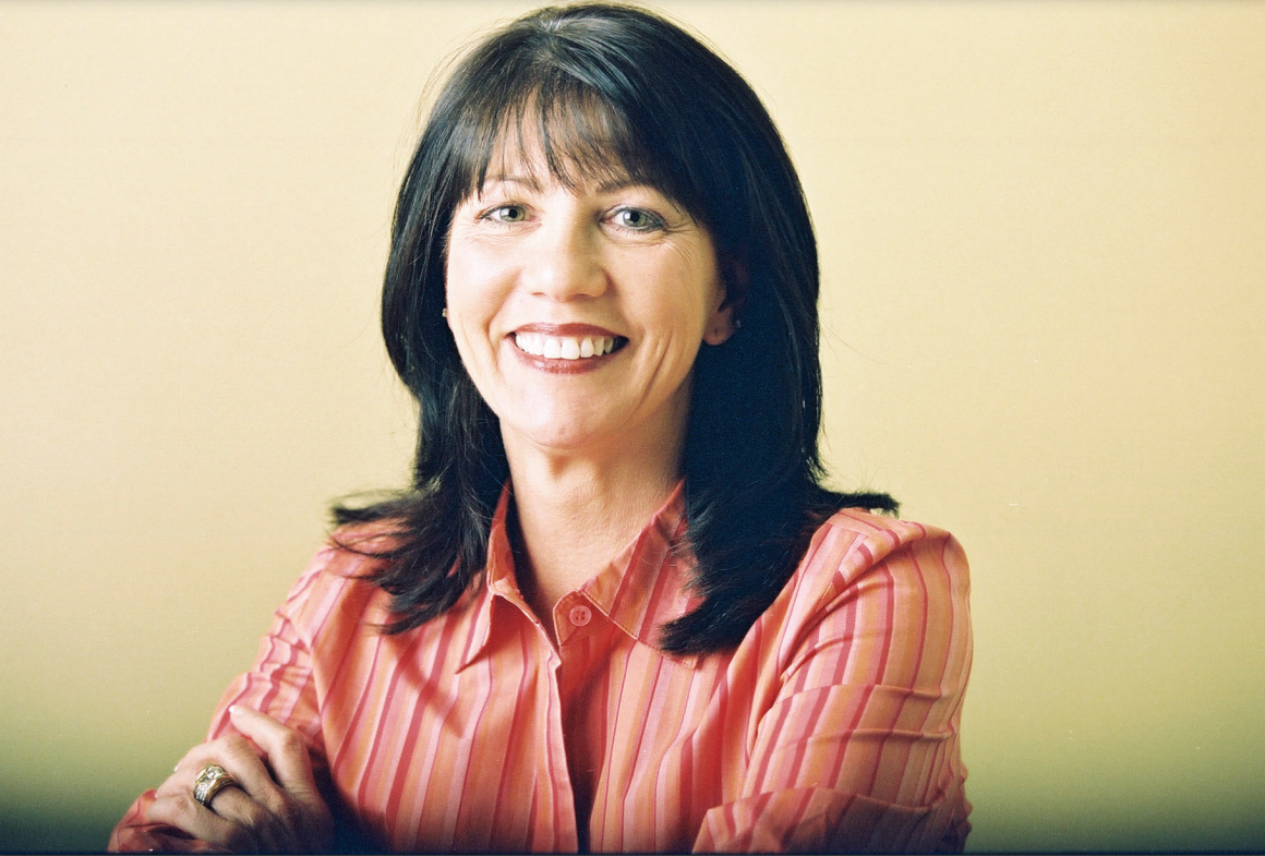 Woman with dark hair, smiling, wearing a pink striped shirt, arms crossed, against a yellow background.