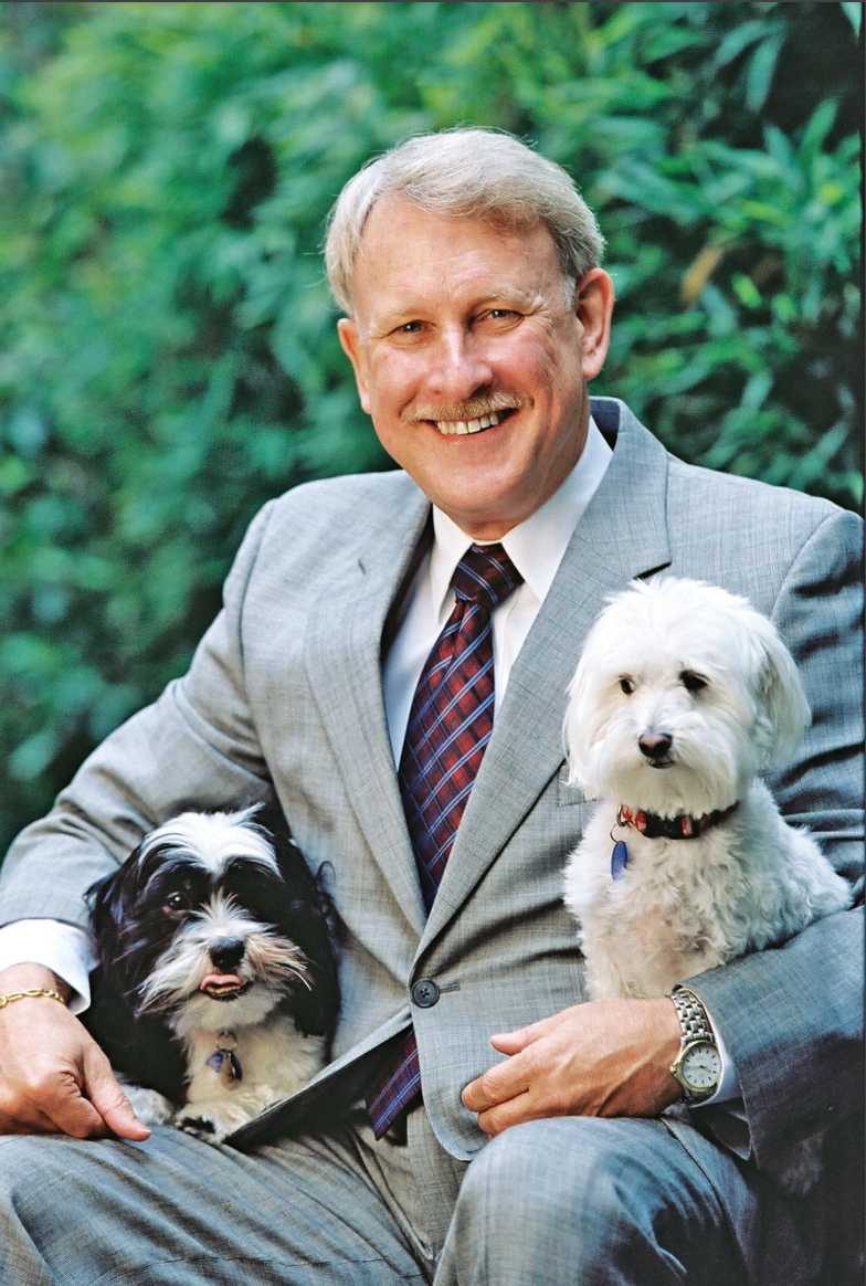 Man in a suit smiles, holding two small dogs in his lap, outdoor setting.