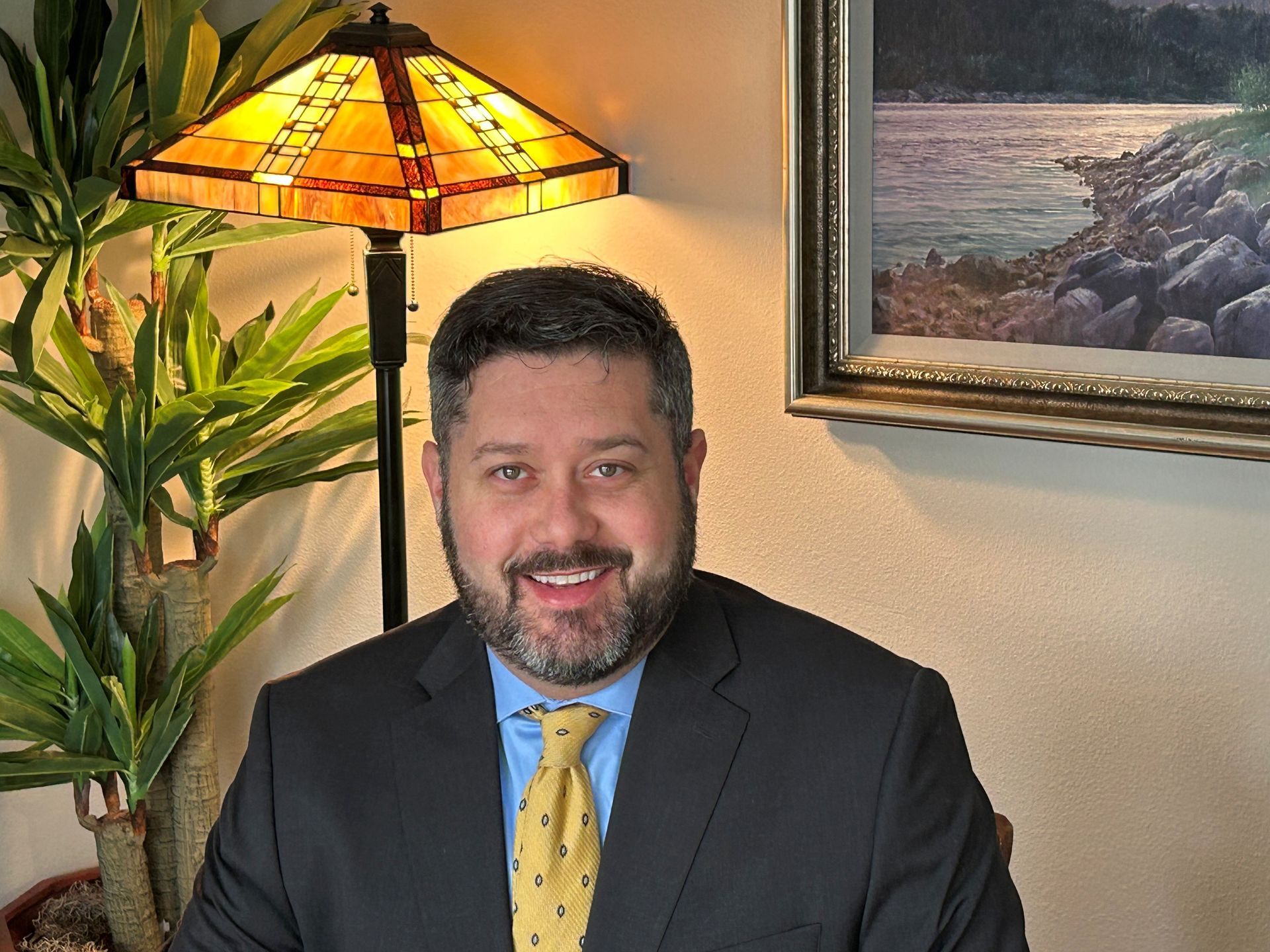 Man in suit smiling, seated near a lit lamp and painting.