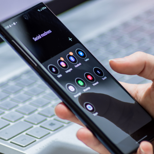 Person holding a black smartphone displaying social media app icons, the screen reflecting a laptop and surroundings.