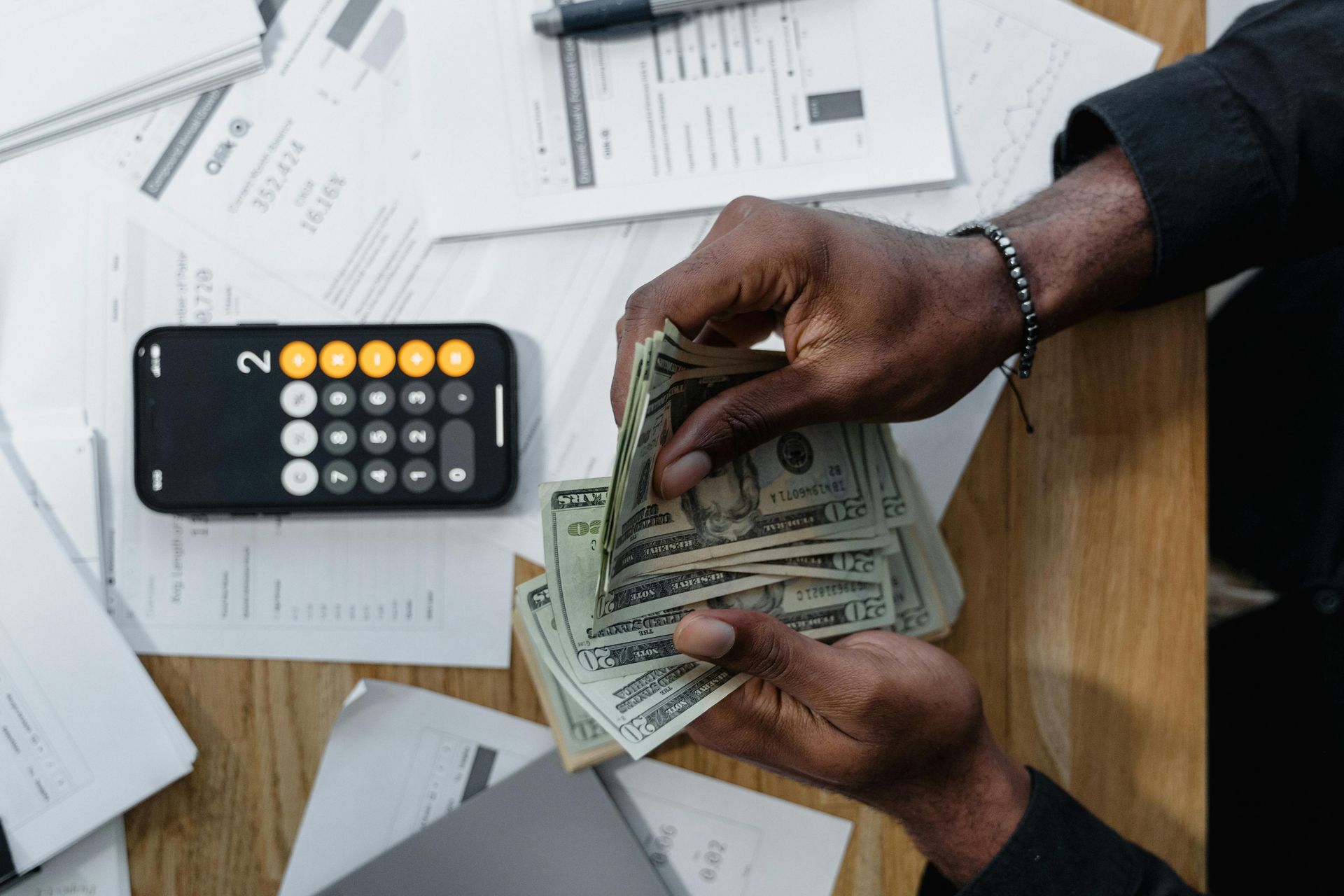 Hands counting US dollar bills on a desk with a calculator and documents.