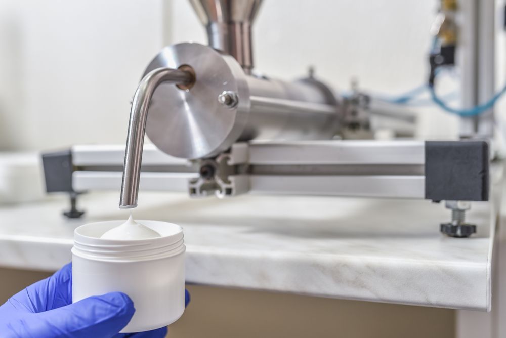A Machine Dispensing White Cream Into a White Plastic Jar Held by a Gloved Hand in a Lab Setting — C & D Manufacturing In Caboolture, QLD