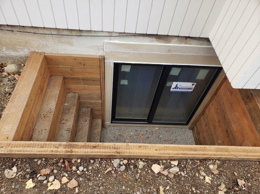 Basement window well with a black sliding window and wooden retaining walls under a house siding