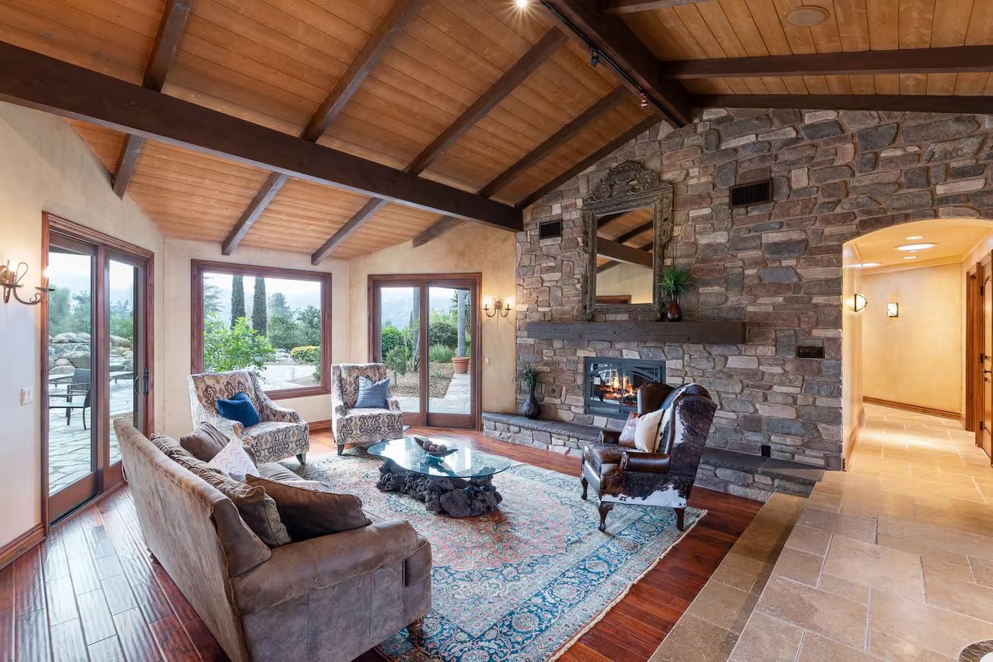 Living room with stone fireplace, wooden ceiling, and large windows overlooking a patio.