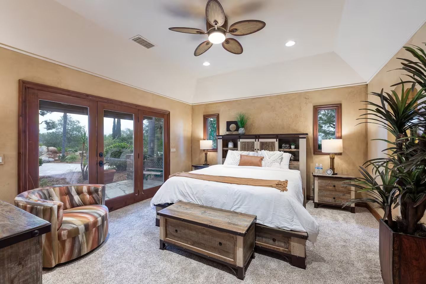 Bedroom with a bed, seating area, and outdoor access through large doors; neutral tones and a ceiling fan.