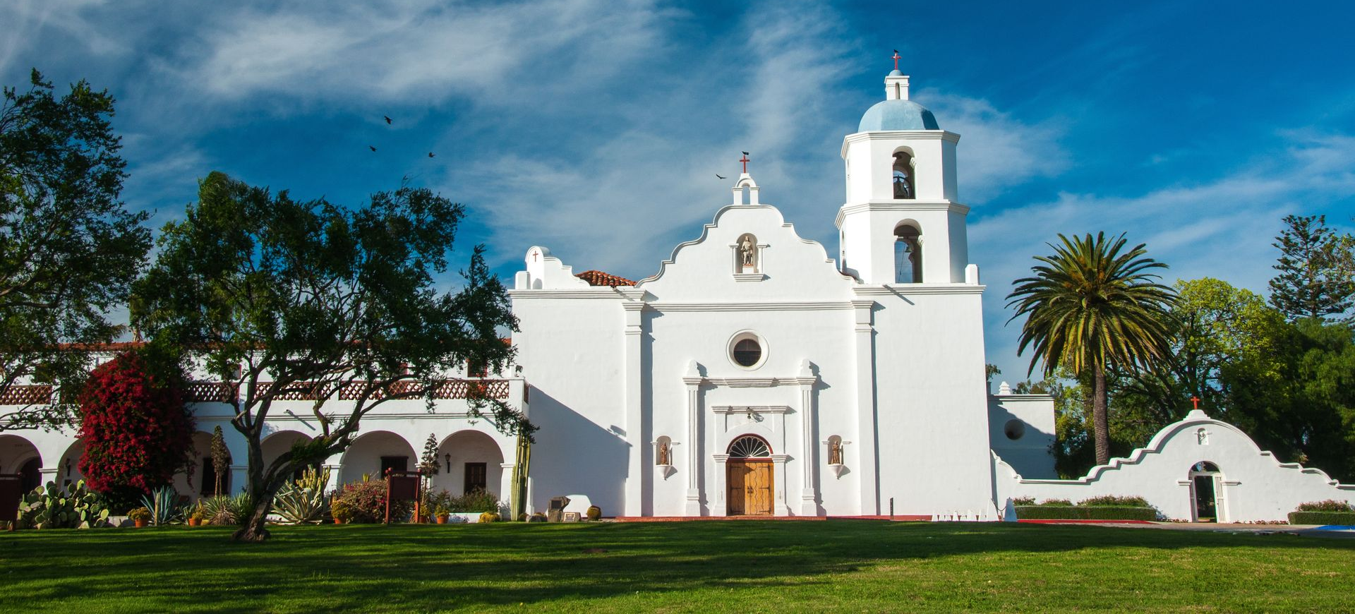 White mission-style church with bell tower and palm trees on a sunny lawn