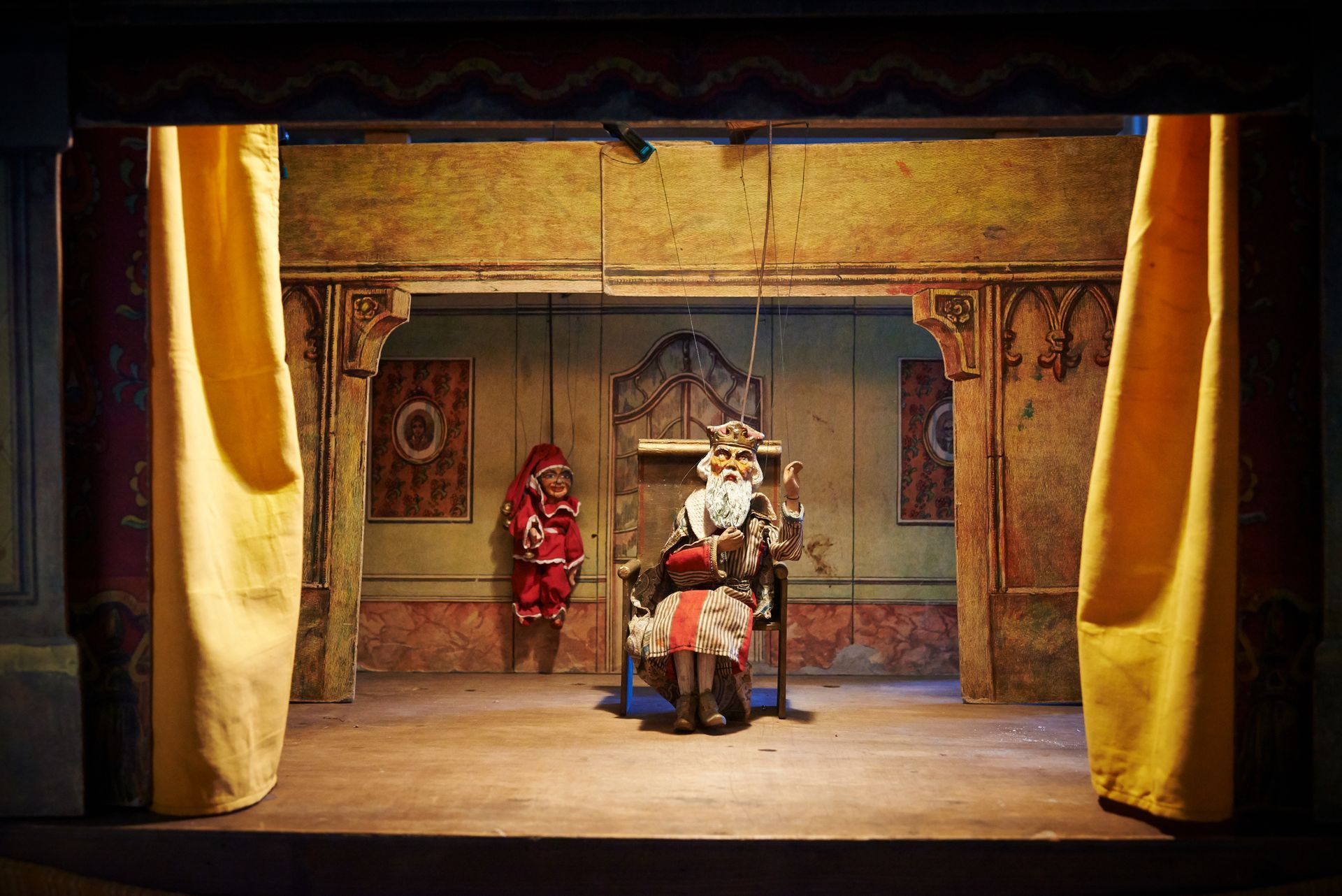 Three performers on a stage set with gold curtains and a painted backdrop, one seated center and two standing nearby