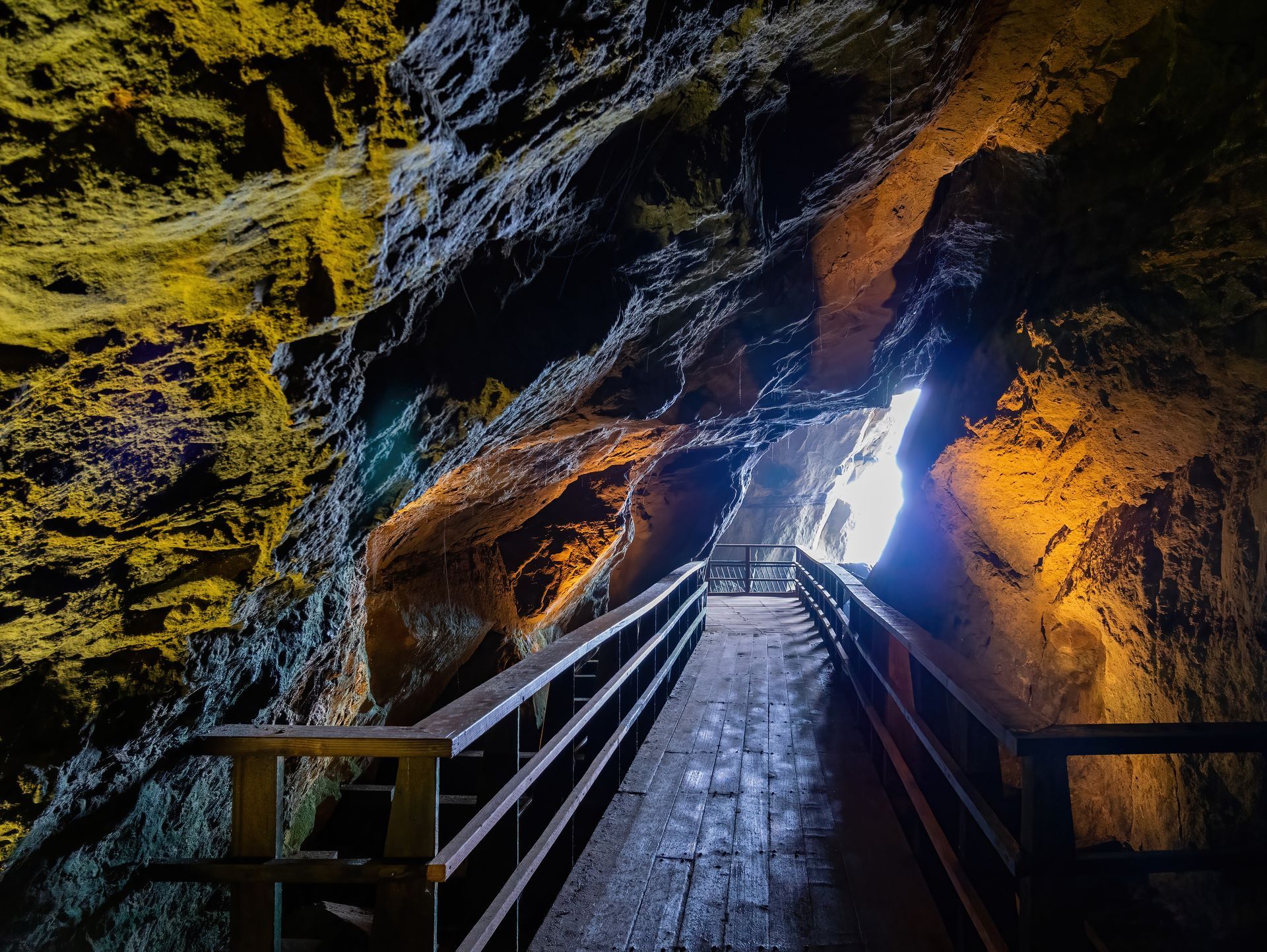 Wooden walkway inside a dim cave leading toward a bright opening, lit by warm cave walls.