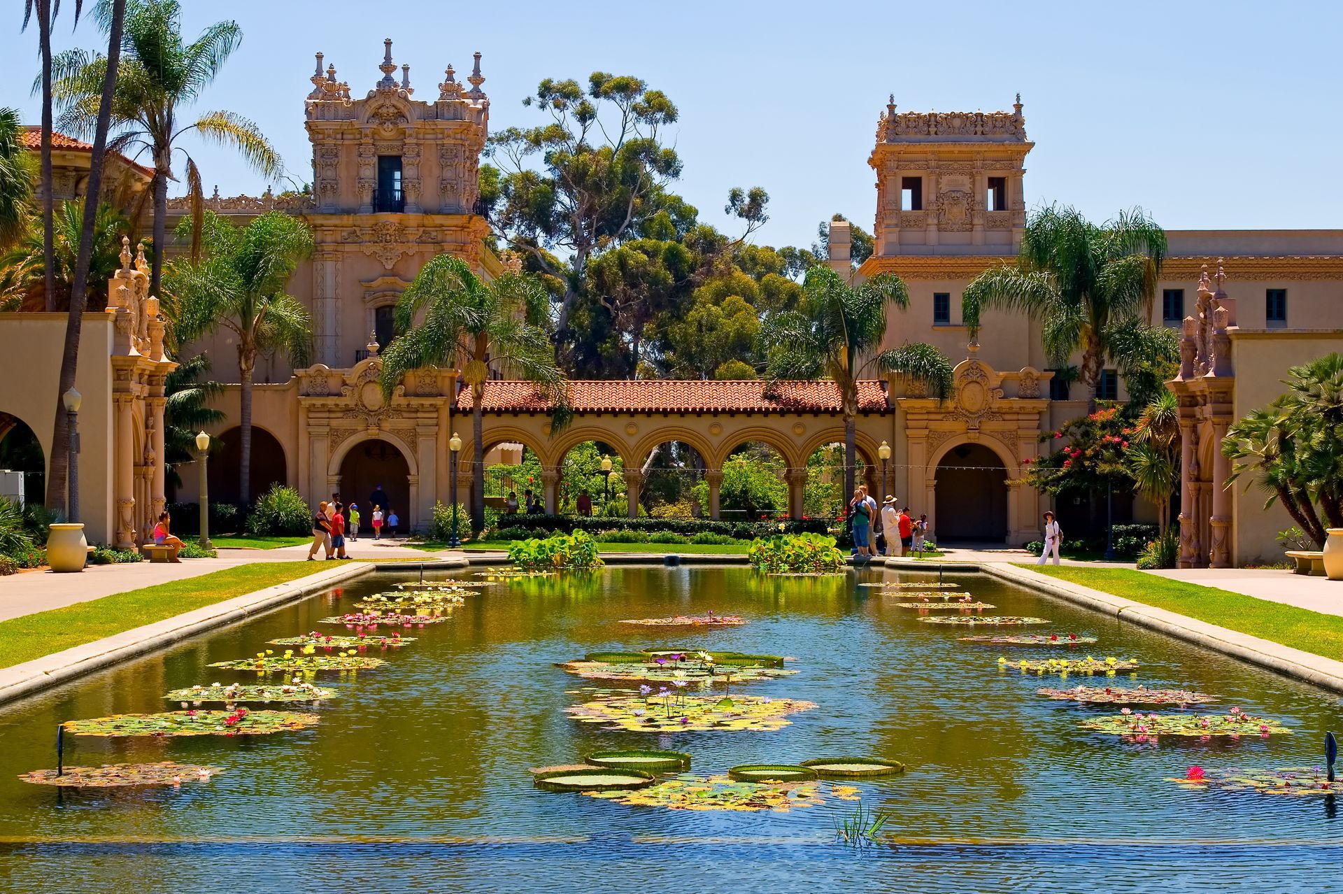 Historic courtyard with reflecting pool, lily pads, arches, and ornate towers under a bright blue sky