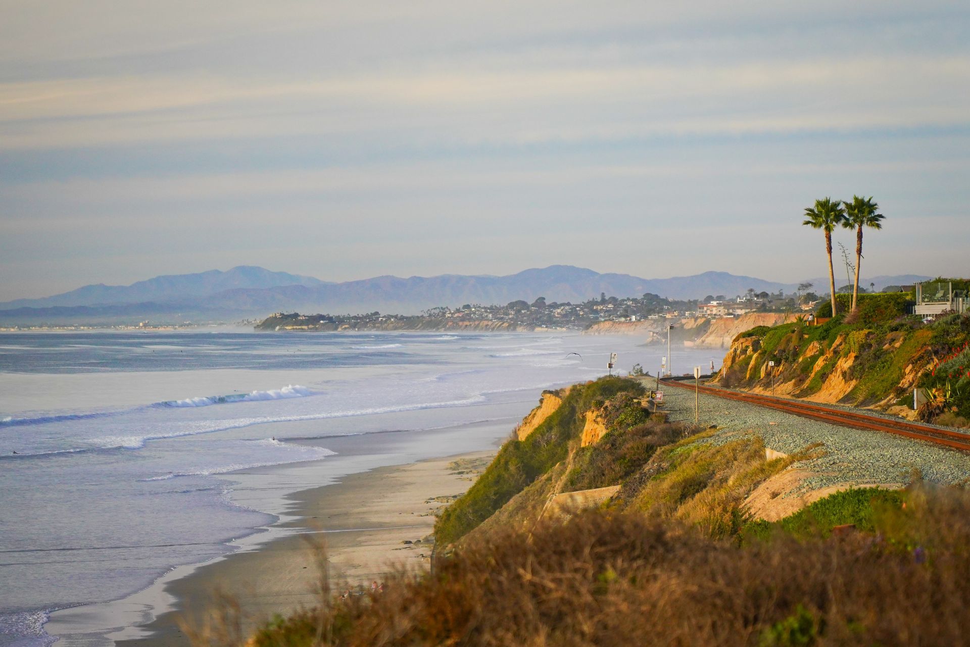 Cliffside coastal road overlooking a sandy beach and calm ocean at sunset, with distant hills and palm trees