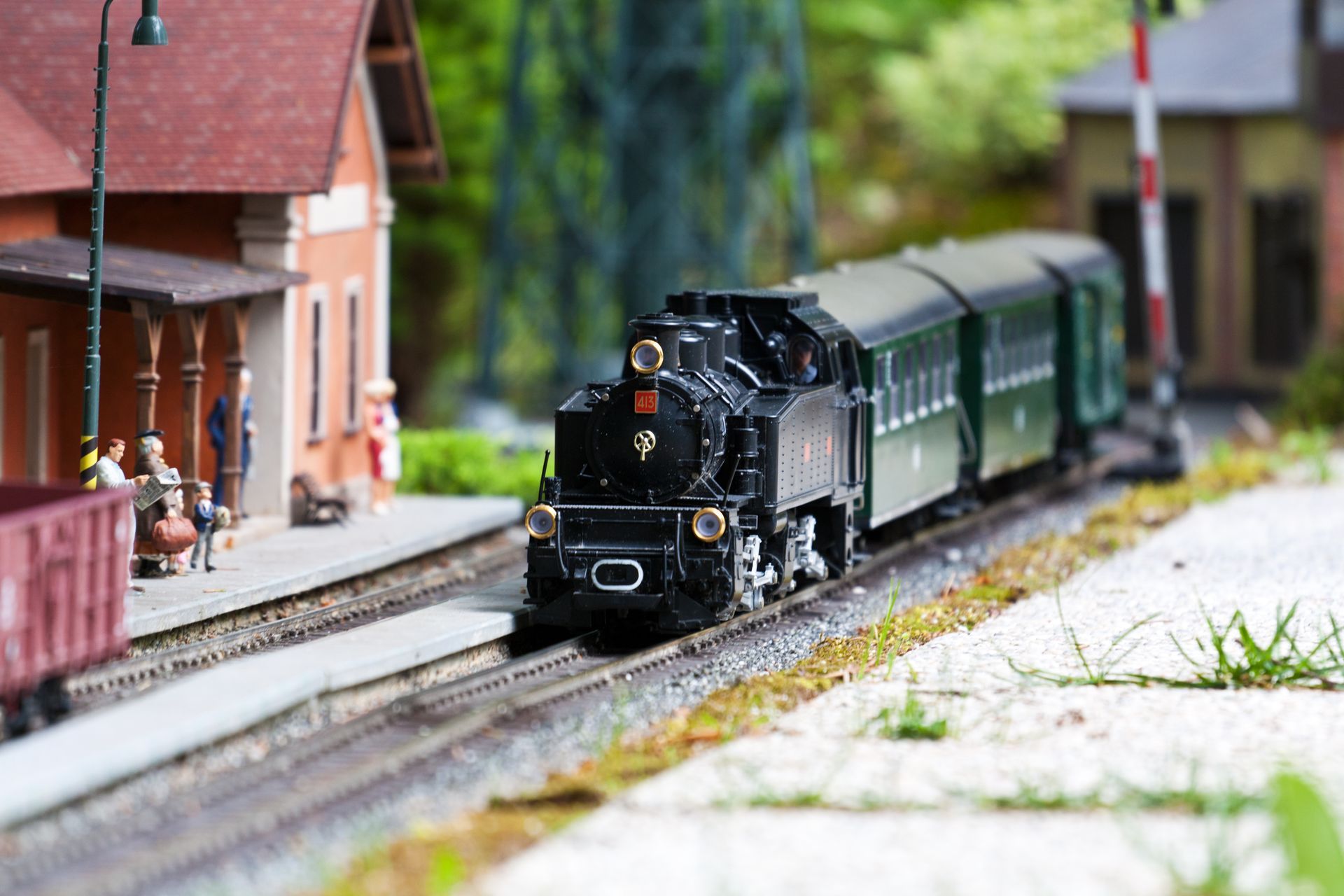 Black model train on miniature station tracks beside a red-brick platform and small figures