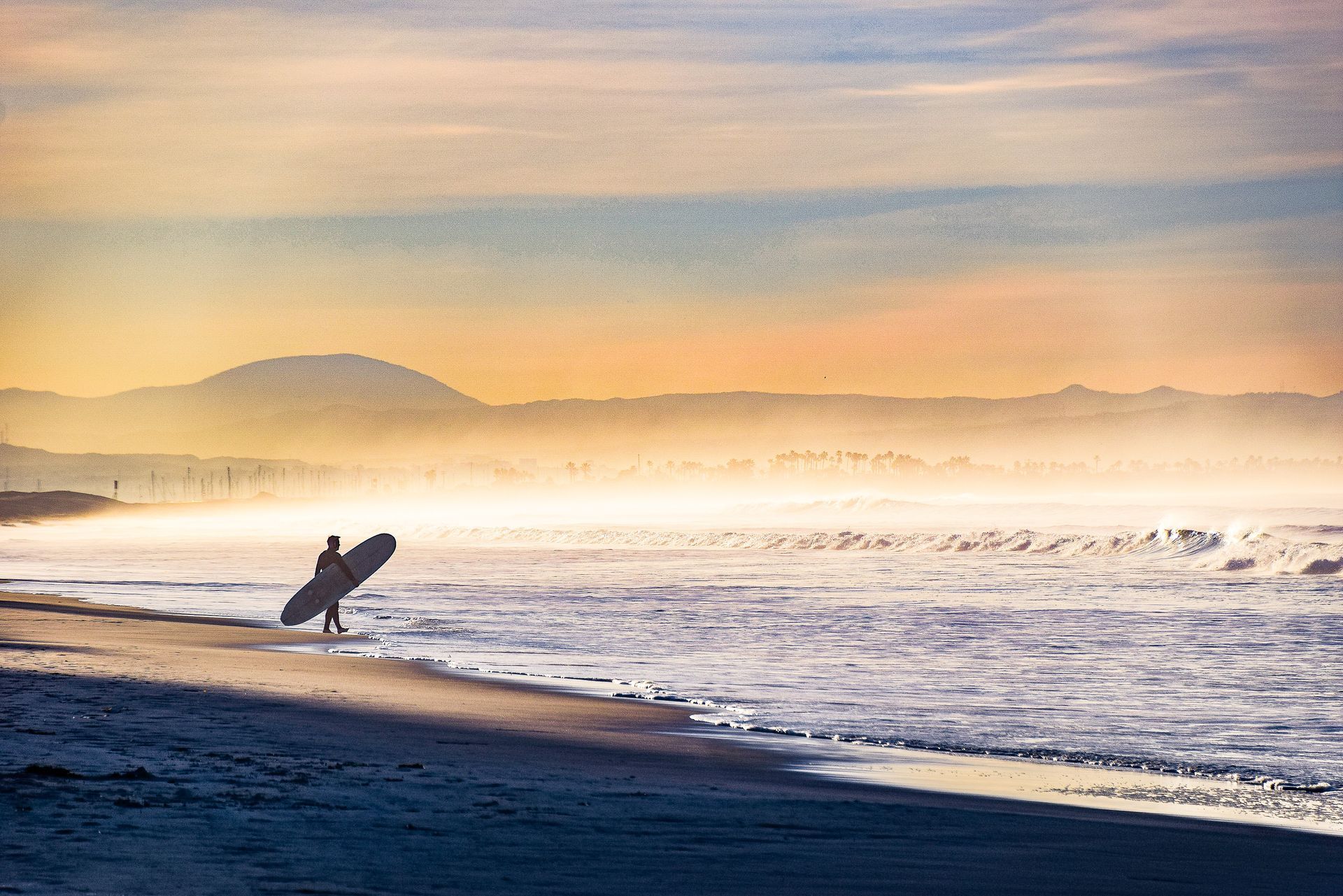 Surfer carrying a board along a misty beach at sunrise, with waves and distant hills.