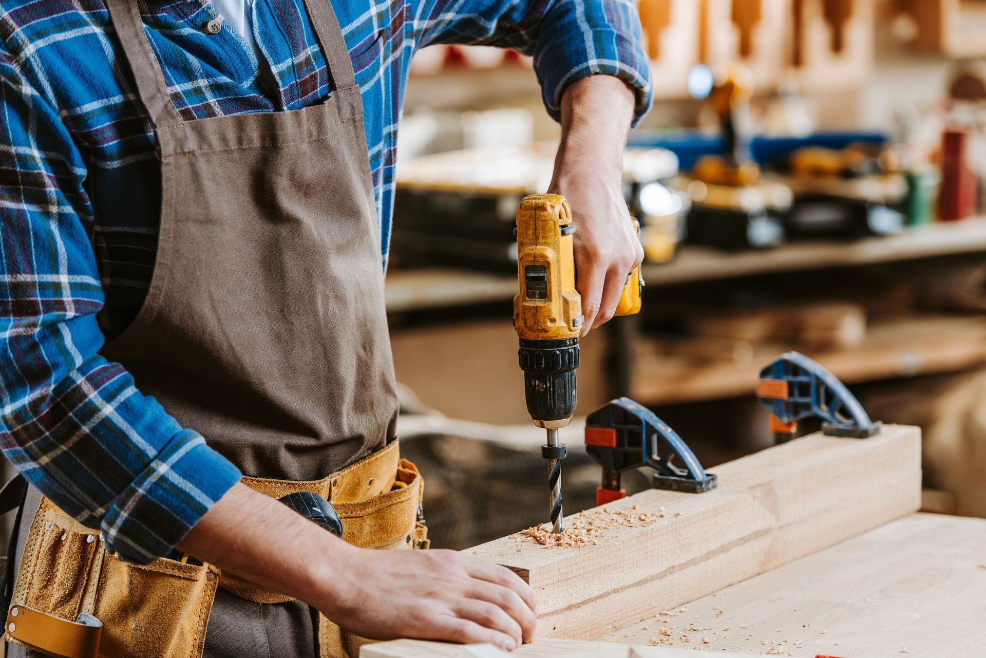 Un uomo con indosso una camicia a quadri e un grembiule sta forando una trave di legno in un'officina.