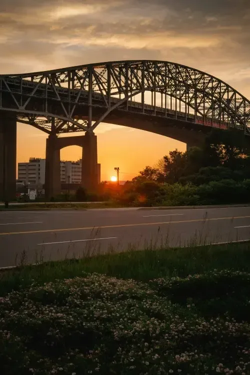 Steel bridge silhouetted at sunset over a highway, with orange sky and city buildings in the distance