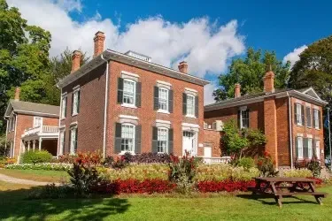 Red brick manor house with white trim, green lawn, flowerbeds, and a picnic table on a sunny day