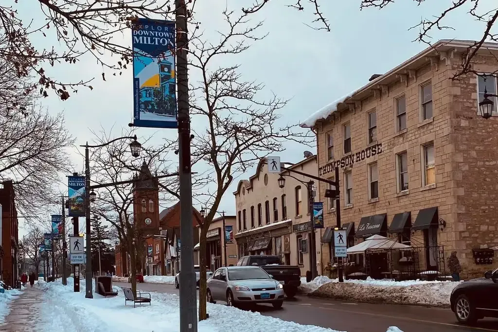 Snowy downtown street with parked cars, leafless trees, and stone storefronts under a cloudy sky