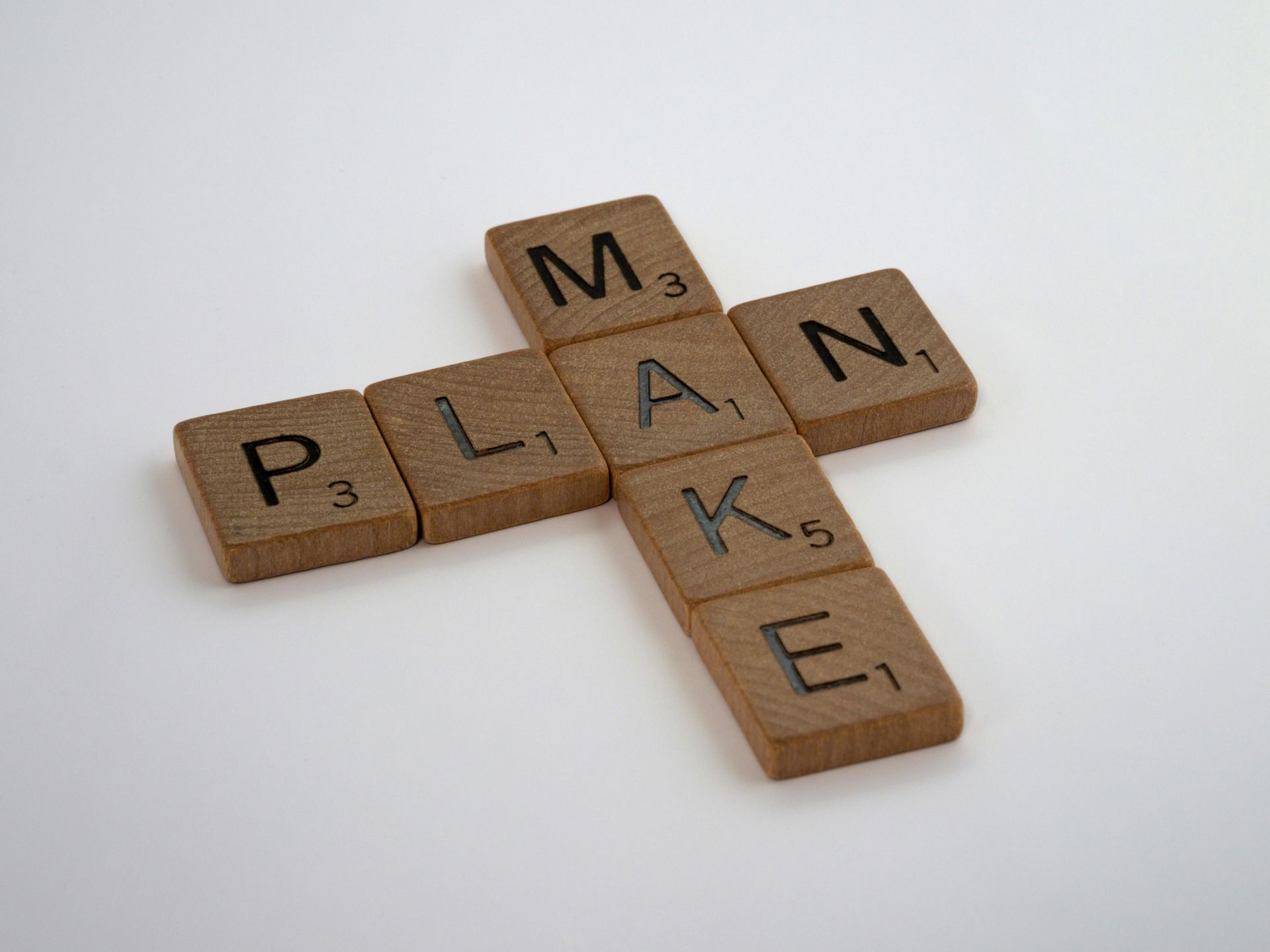 Scrabble tiles spelling “MAKE PLAN” arranged in a cross on a white background