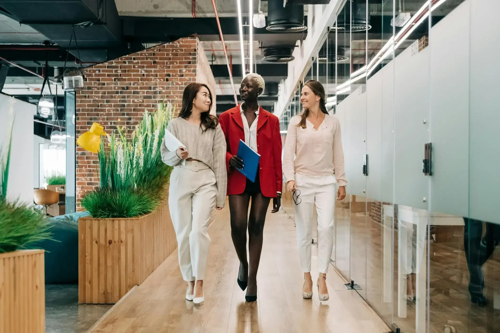 Three people walking through a modern office hallway, surrounded by plants and glass walls.