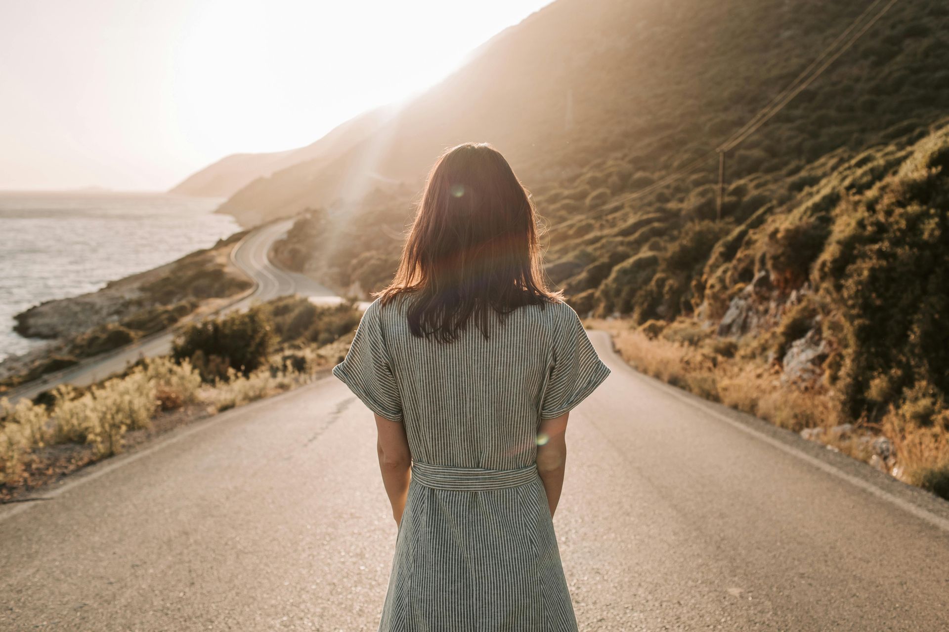 Person standing on a sunlit coastal road beside hills and ocean at sunset