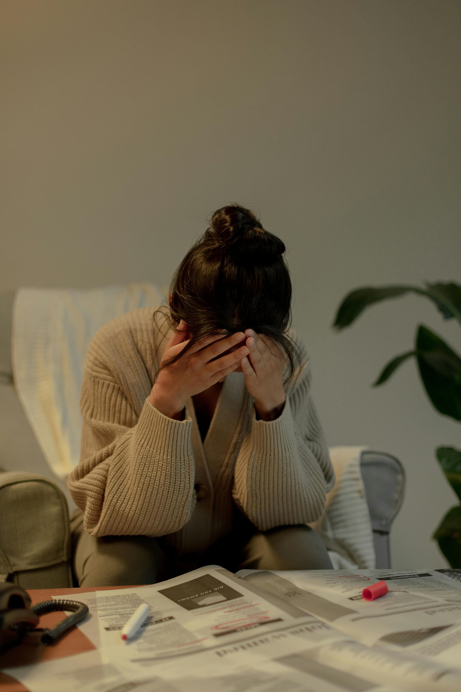 Person sits at a desk with head in hands, surrounded by papers and a phone, looking stressed.