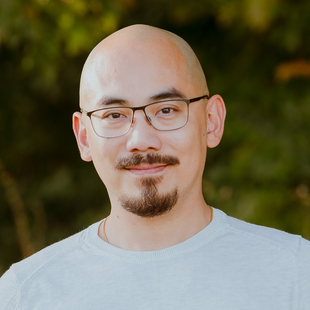 Portrait of a bald man with glasses and a goatee, wearing a light shirt against a blurred outdoor background