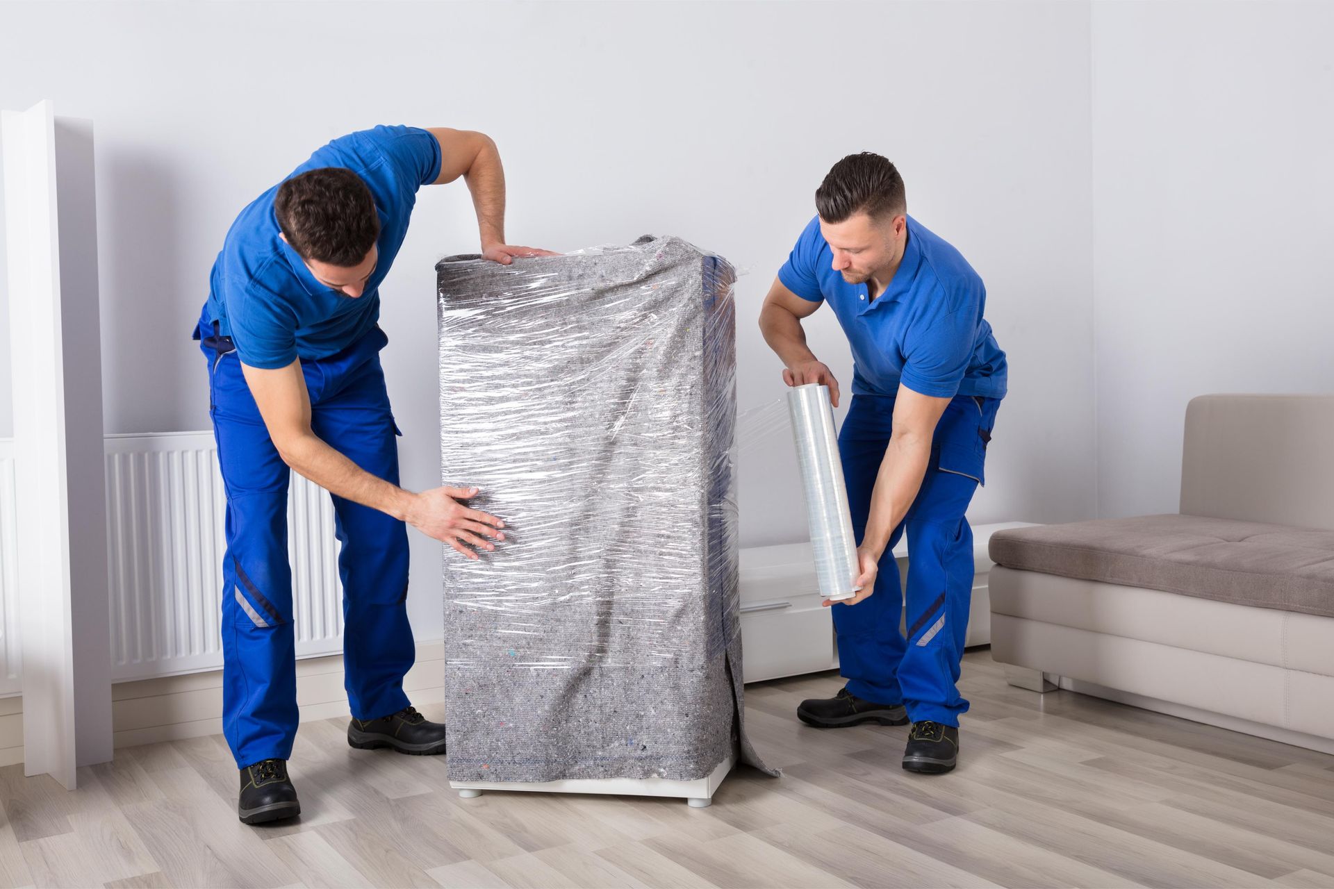 Two movers in blue uniforms wrapping furniture with plastic wrap indoors.