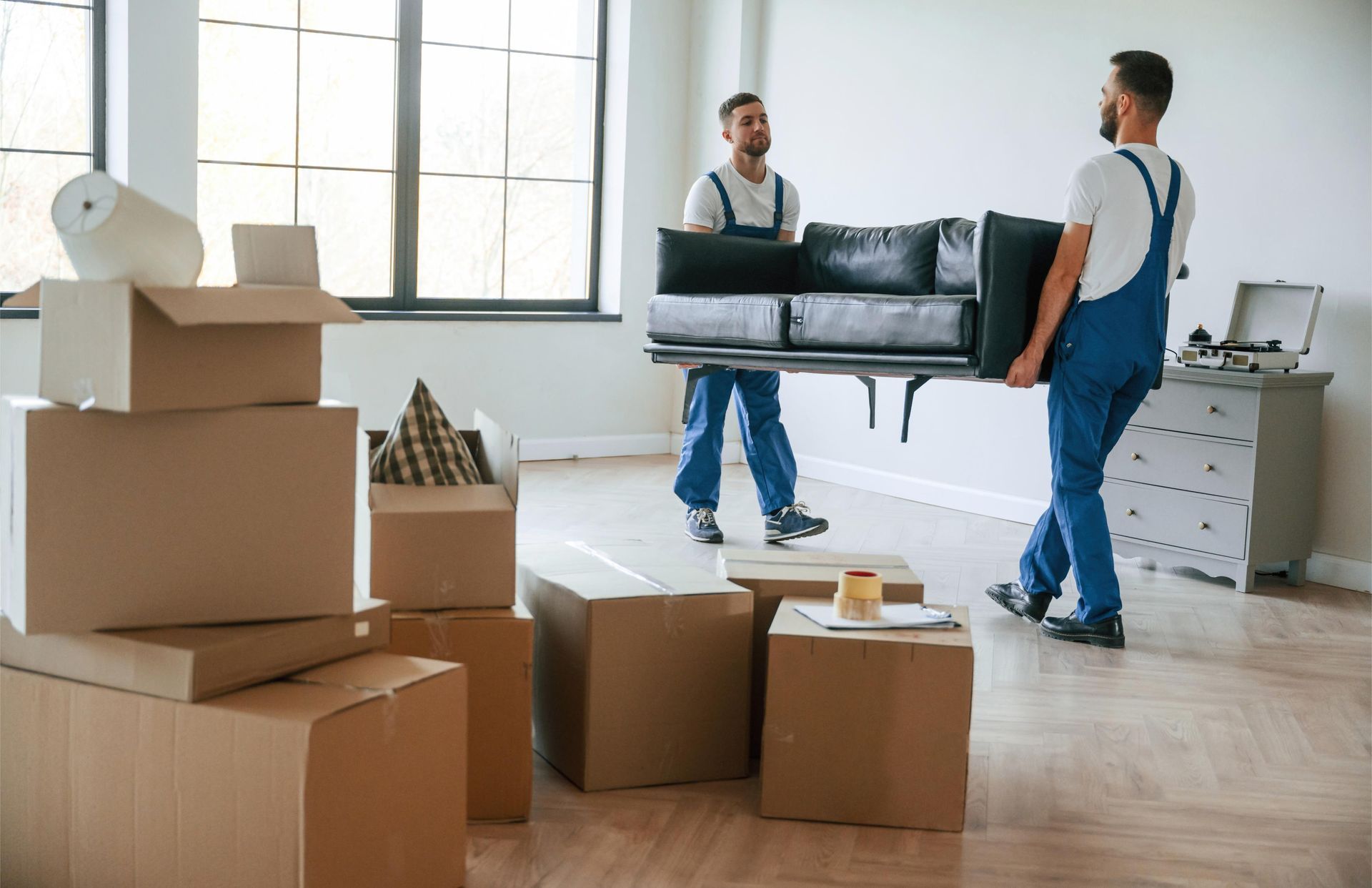 Two movers in blue overalls carrying a black sofa into a new home with packed boxes.