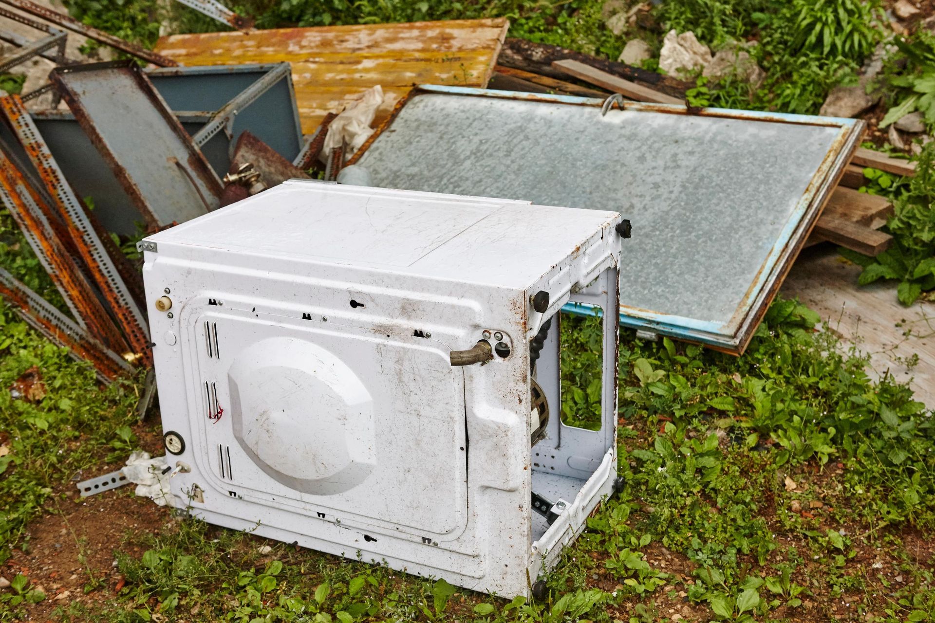 Discarded white washing machine frame with broken windows, outdoors on grass.