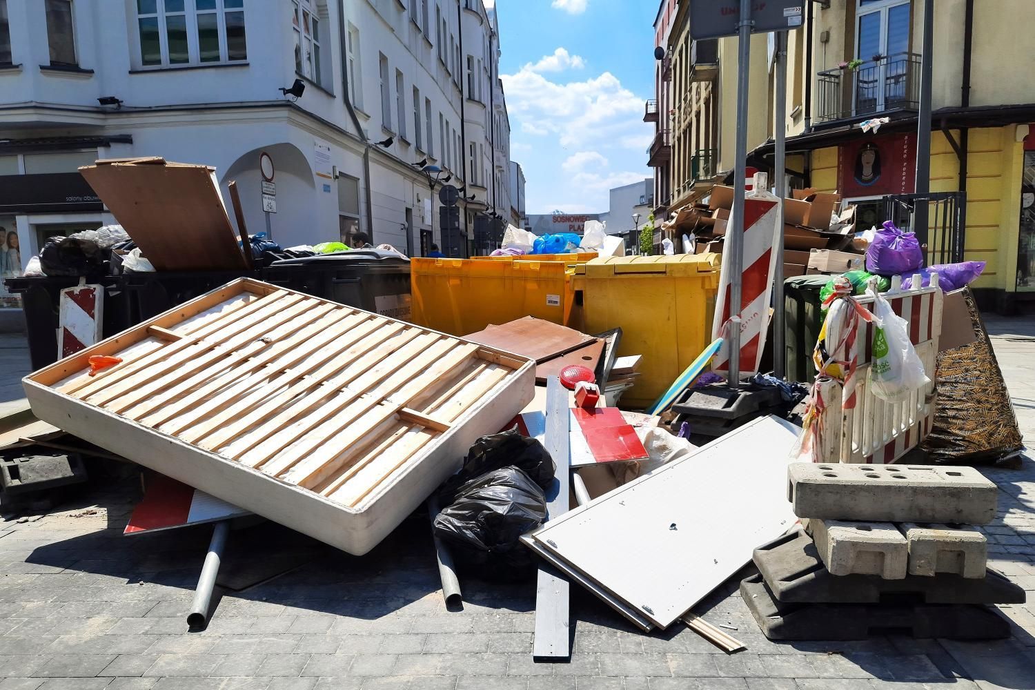 Street scene with trash overflowing from bins, including a bed frame and construction debris.