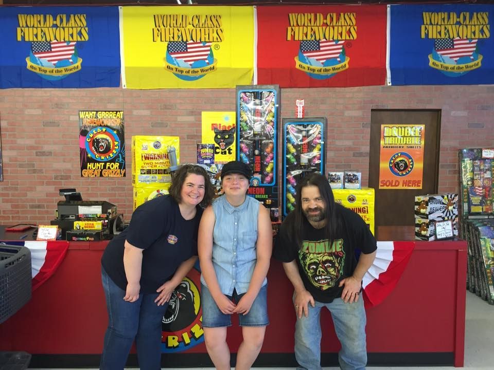 Three people are posing for a picture in front of a world class fireworks sign