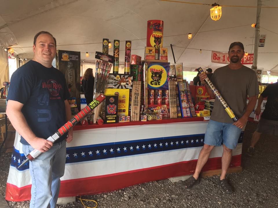 Two men are standing in front of a table full of fireworks.