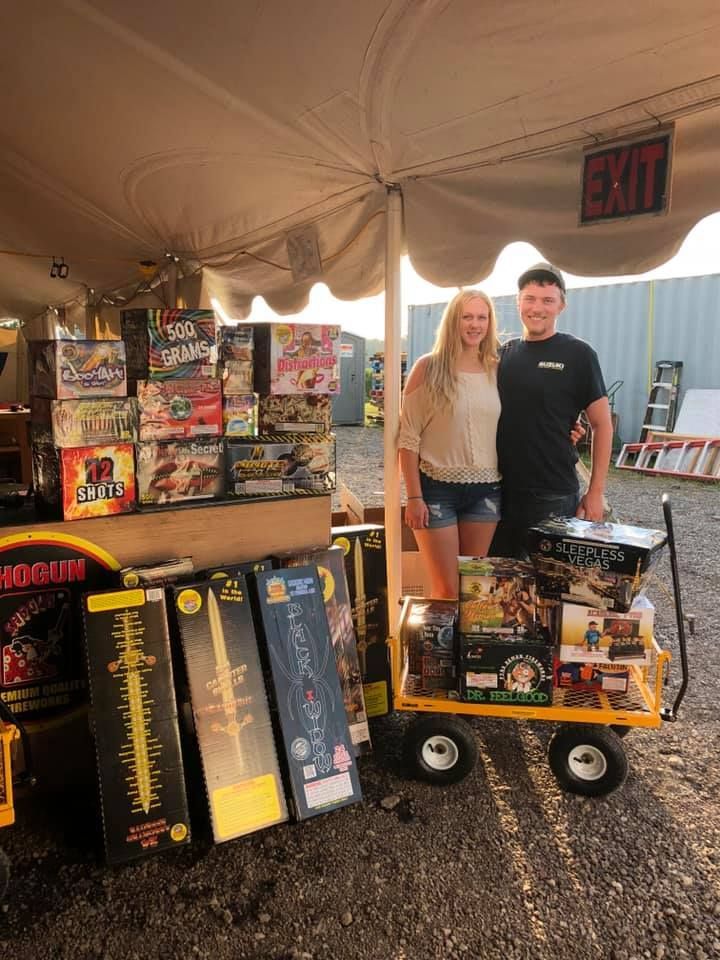 A man and a woman are standing in front of a cart filled with fireworks.