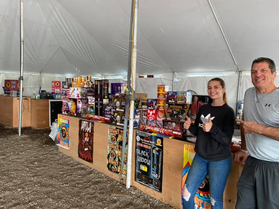 A man and a woman are standing in front of a fireworks stand under a tent.