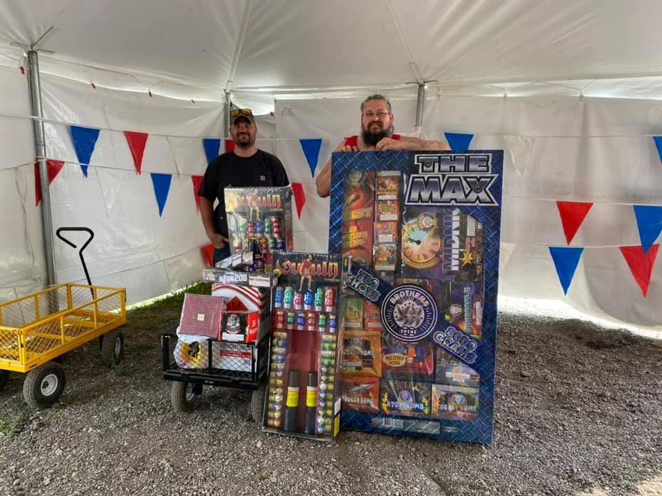 Two men are standing under a tent holding a large box of fireworks.