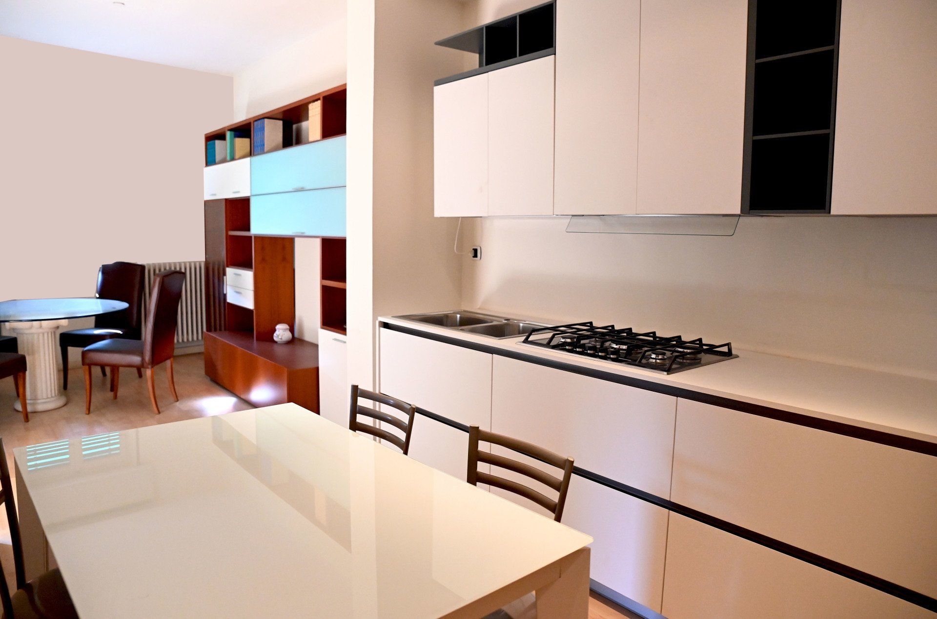 Cucina moderna con mobili bianchi, piano cottura a gas, tavolo da pranzo e scaffalature in un luminoso ambiente aperto.