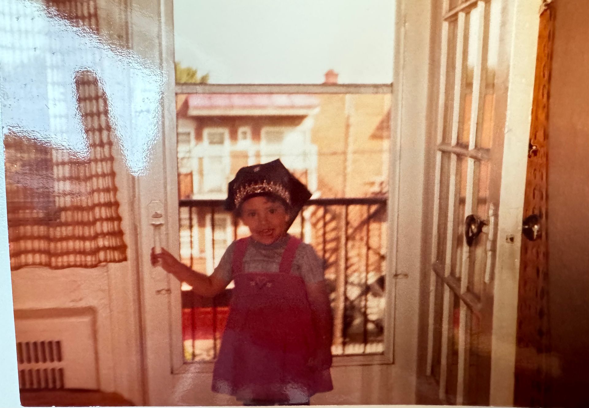 Child in pink overalls and a hat standing in an open doorway, looking at the camera. Buildings visible outside.