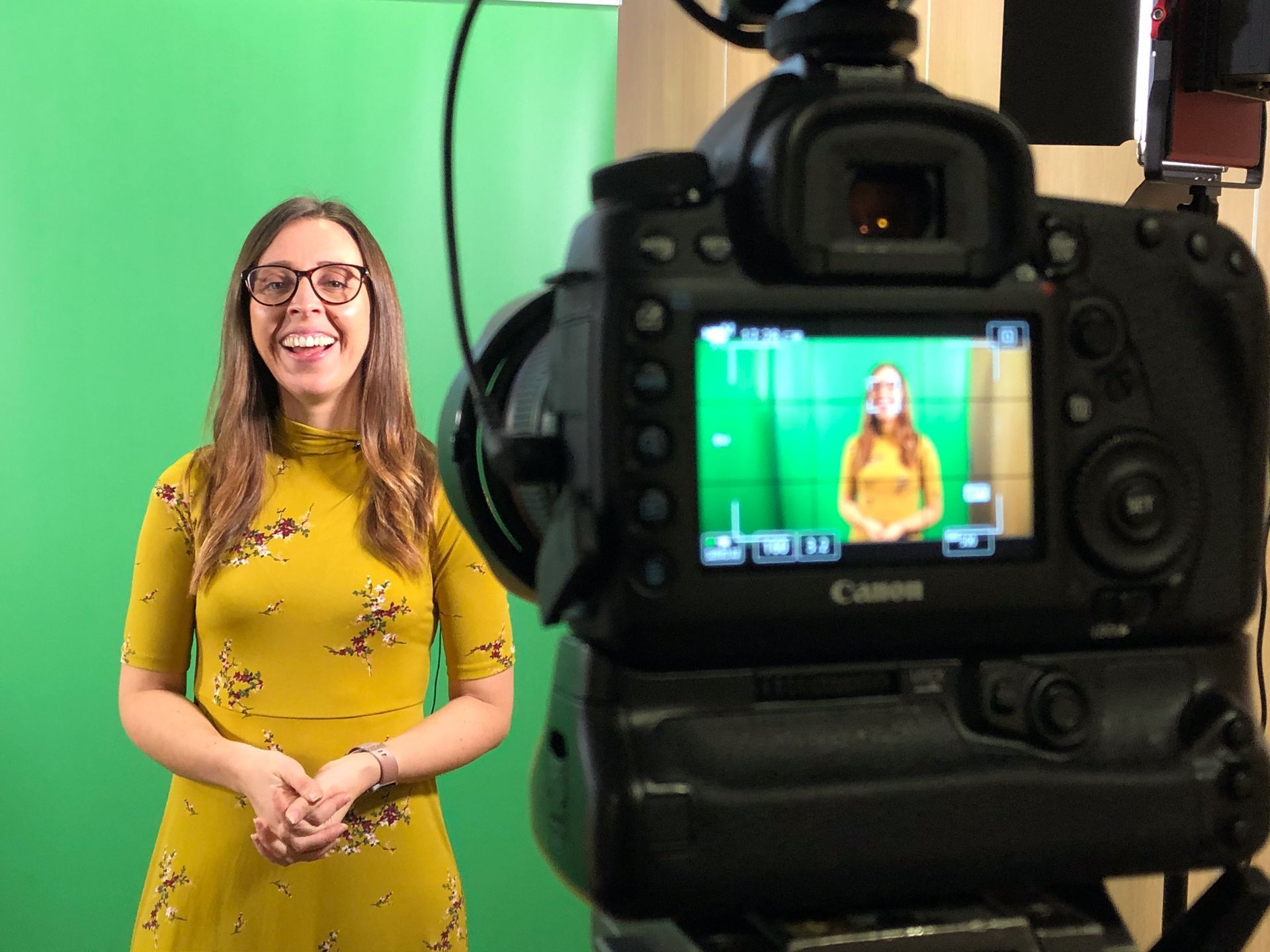 A person in a yellow floral dress standing in front of a green screen, viewed from behind a professional camera.