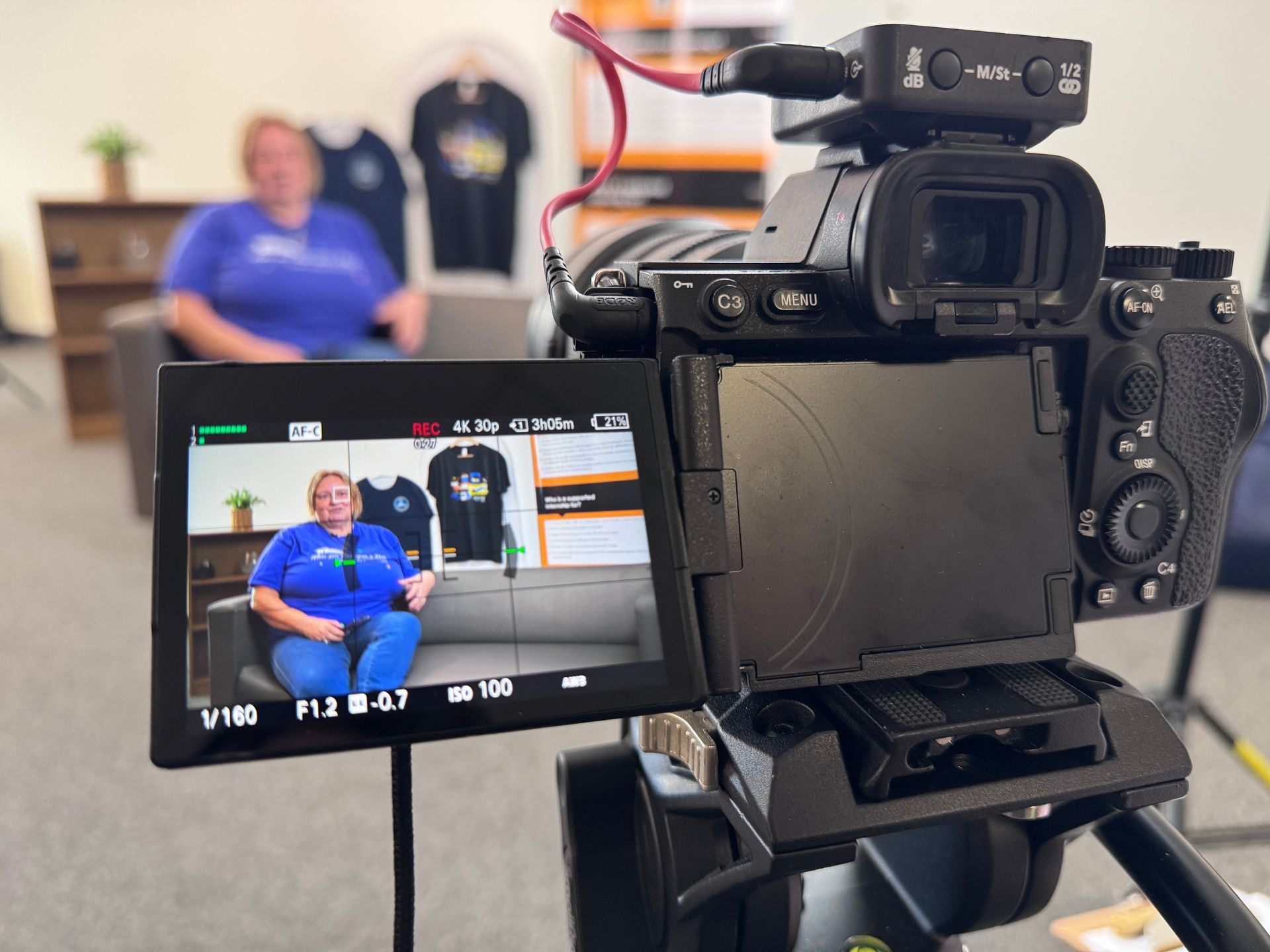 A camera filming a person sitting on a sofa in a studio setting, shown through the camera’s flip-out viewfinder display.