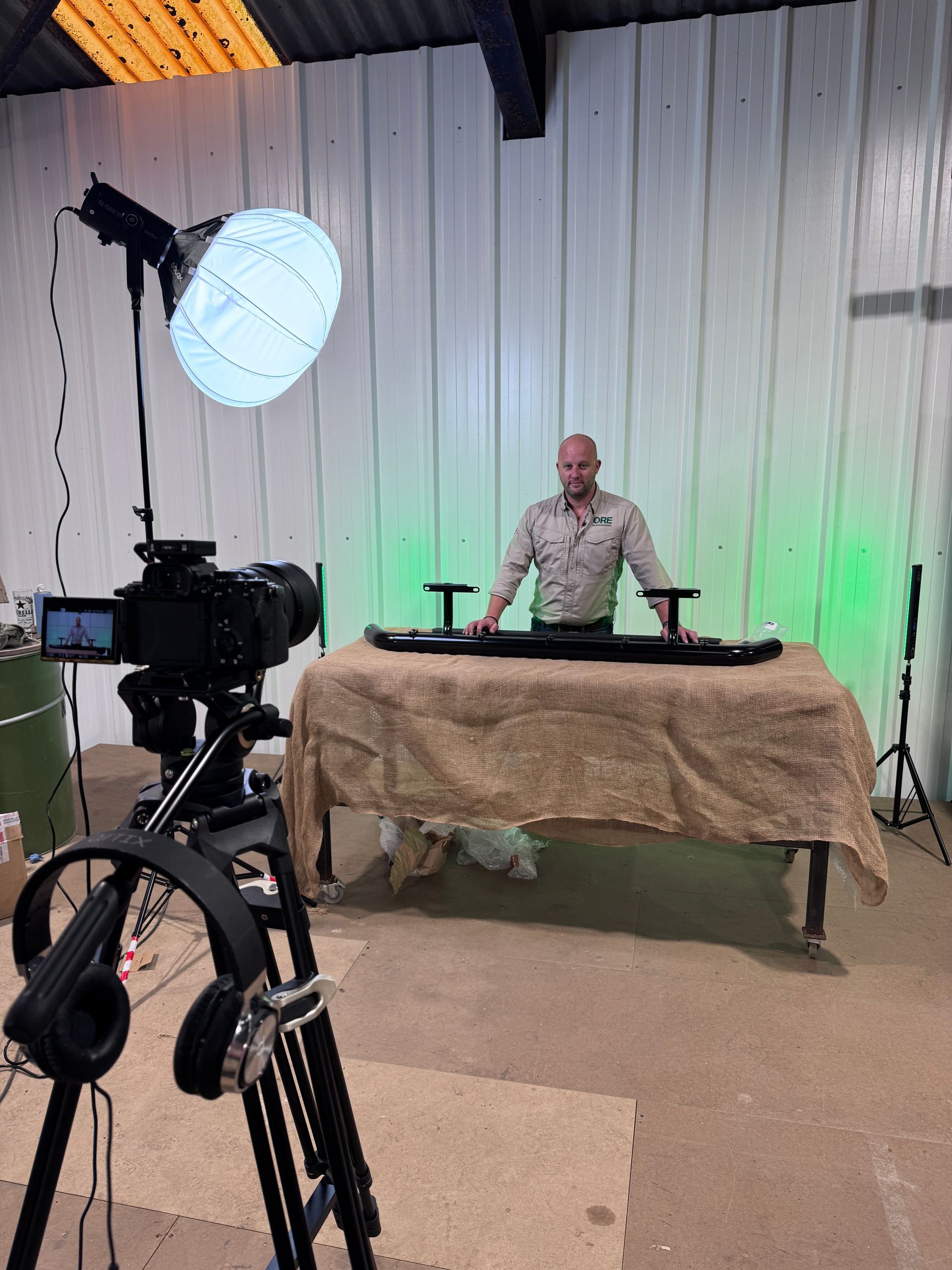 A person stands behind a table with a black object in a workshop, facing a professional camera and lighting setup.