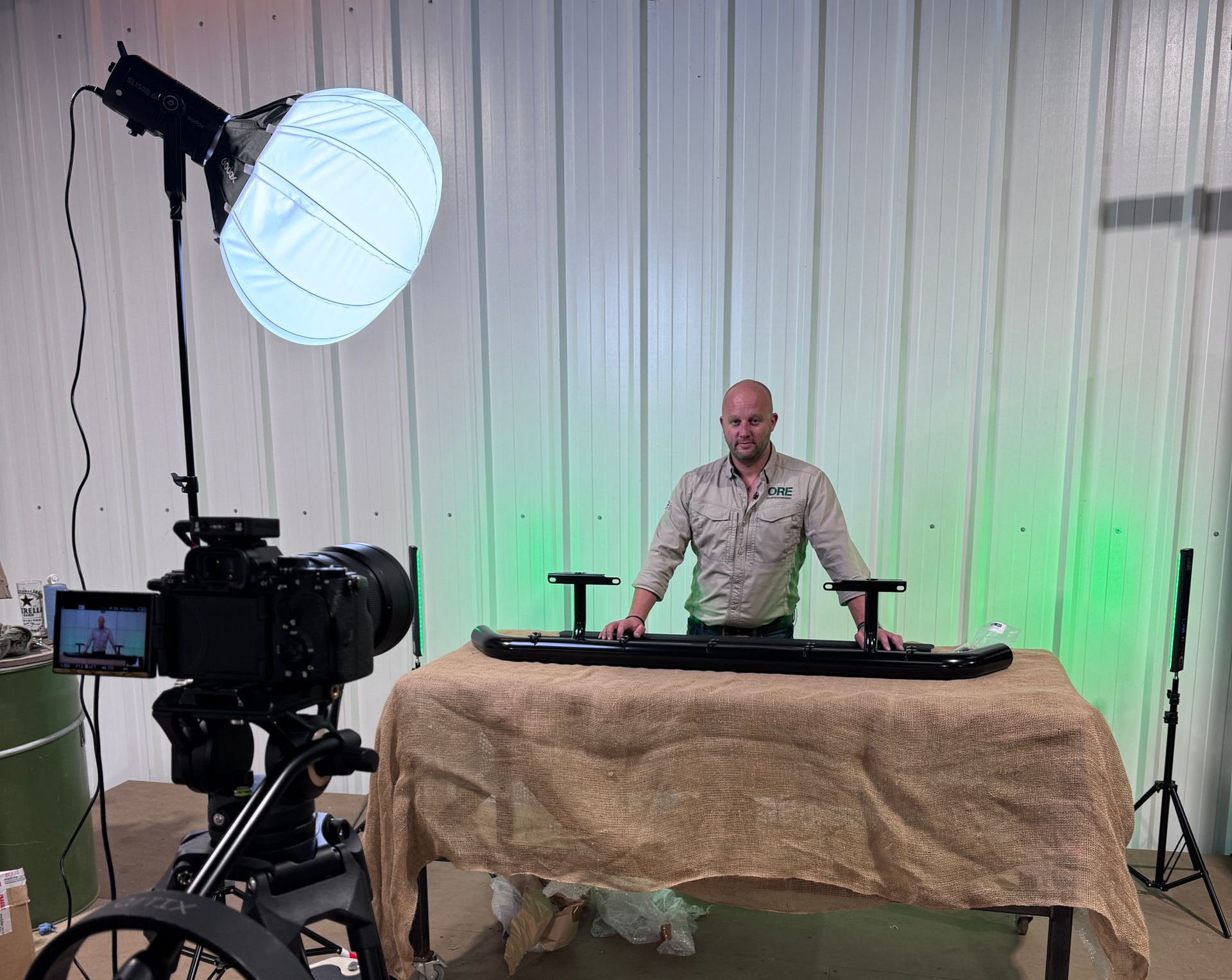 A person stands behind a table with a black bumper assembly in a studio setting with lighting equipment and cameras.