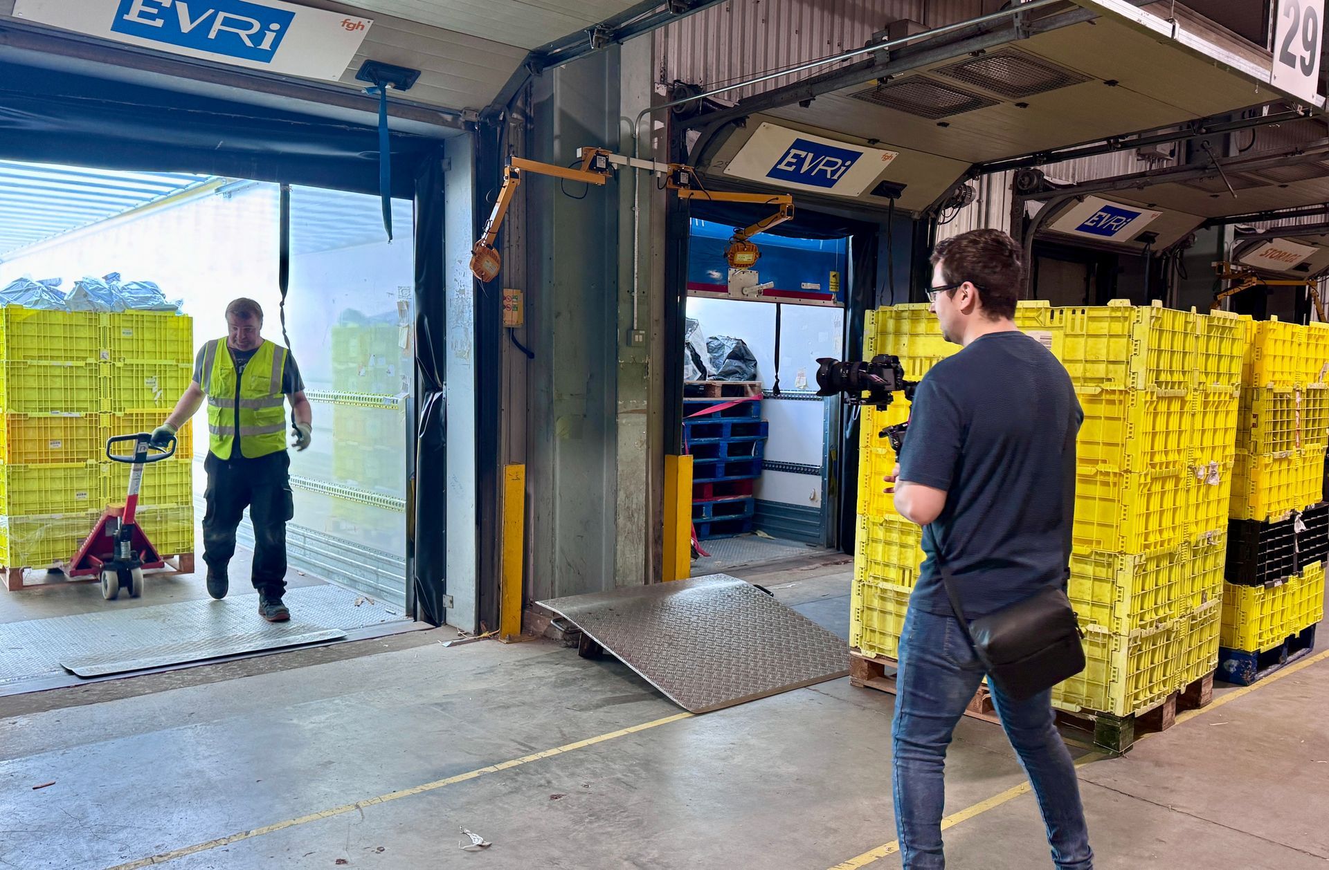 A cameraman records a worker maneuvering a stack of yellow crates using a pallet jack at an Evri distribution center.