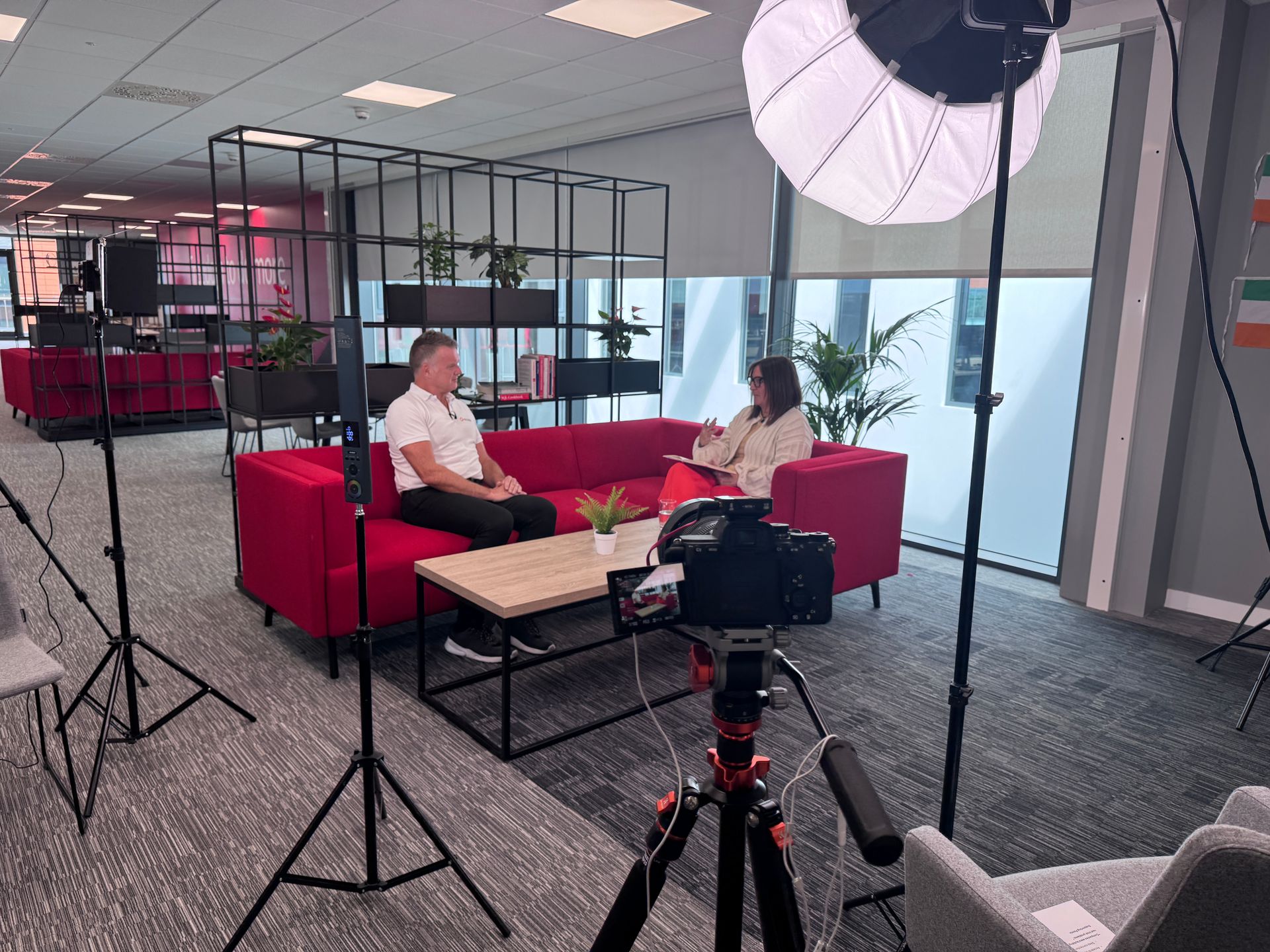 A professional video interview setup in an office with two people sitting on a red sofa, captured by a camera on a tripod.