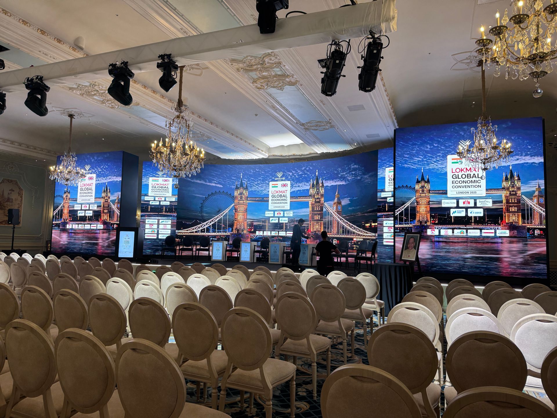 Rows of empty chairs facing a large stage with a screen displaying Tower Bridge in an elegant, chandelier-lit ballroom.
